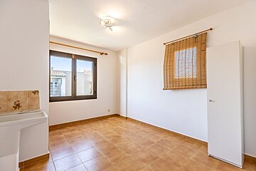 An empty room with white walls, terracotta tile floors, two windows (one with a bamboo blind), a small sink, and a white cabinet.