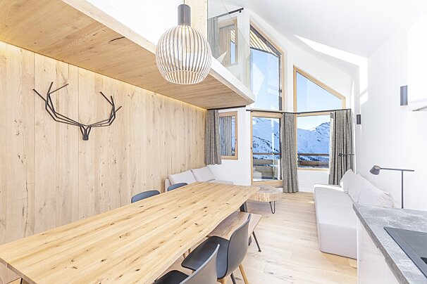 Bright, modern chalet interior with a long wooden dining table, minimalist decor, and large windows overlooking snowy mountains.