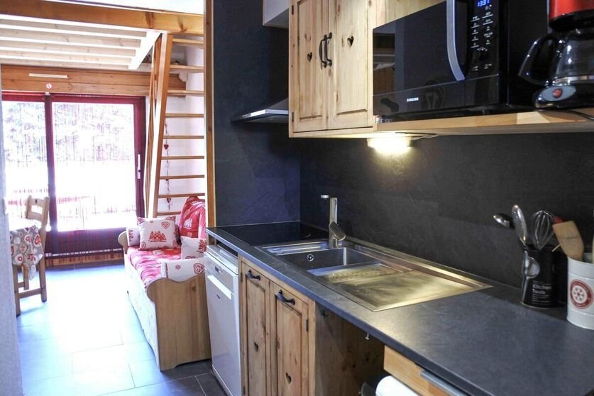 A rustic apartment interior featuring a compact kitchen with wooden cabinets and dark counters, a red-patterned sofa, and a wooden ladder to a loft.