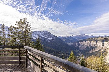 A wooden balcony offers a panoramic view of snow-capped mountains, a deep valley with evergreen forests, and a town under a cloudy blue sky.