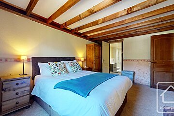 Cozy bedroom with exposed wooden beams, a large bed with a blue throw, floral pillows, bedside tables, and an en-suite bathroom.