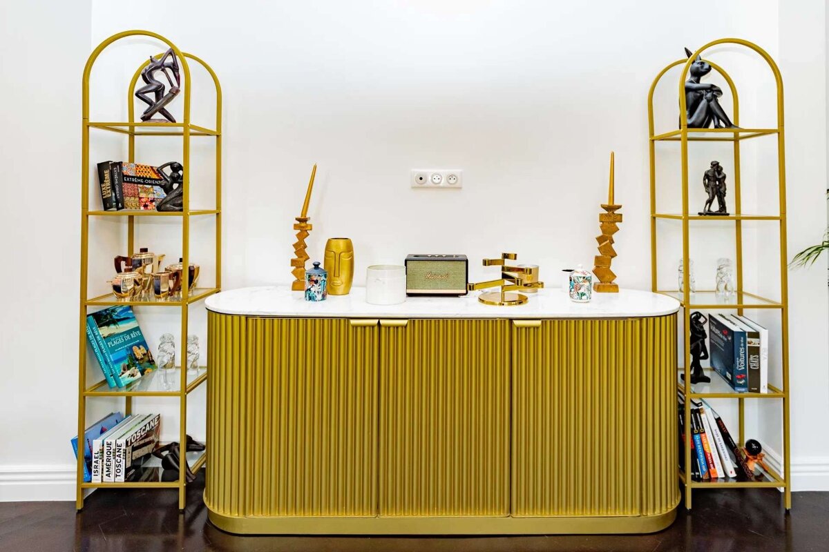 A chic gold fluted cabinet with a white marble top sits between two arched gold shelving units, adorned with books, sculptures, and a speaker against a white wall.