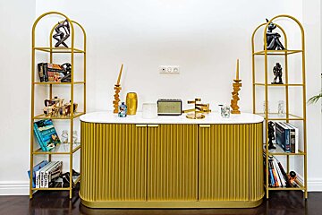 A chic gold fluted cabinet with a white marble top sits between two arched gold shelving units, adorned with books, sculptures, and a speaker against a white wall.