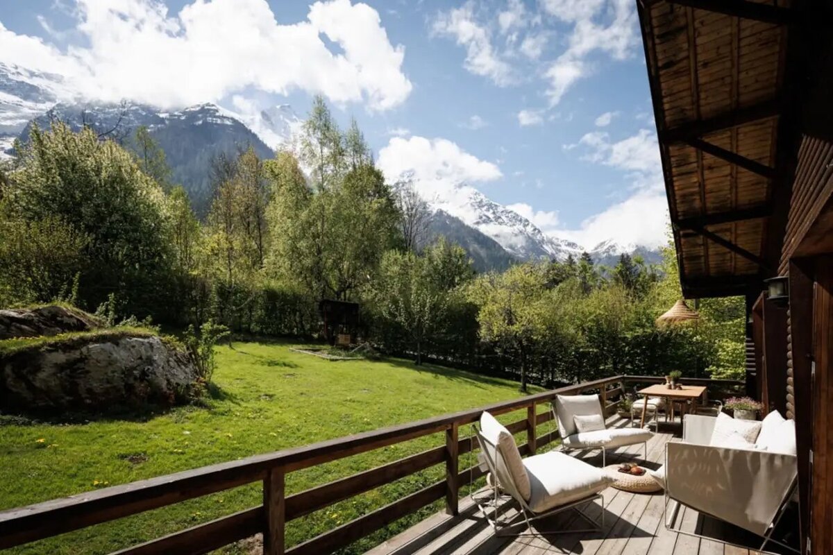 Scenic wooden deck with white lounge chairs overlooking a lush green yard and majestic snow-capped mountains under a bright blue sky.
