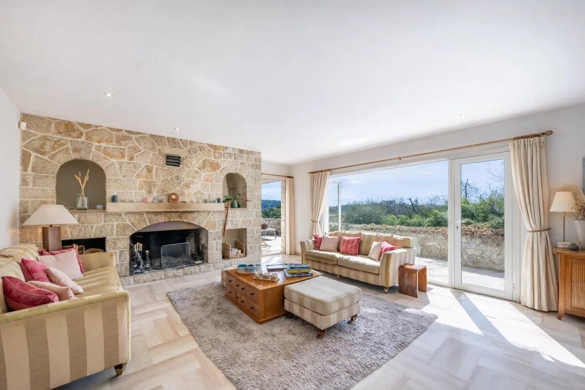 Sunlit living room featuring a rustic stone fireplace, comfortable sofas, and large glass doors opening to a scenic view.