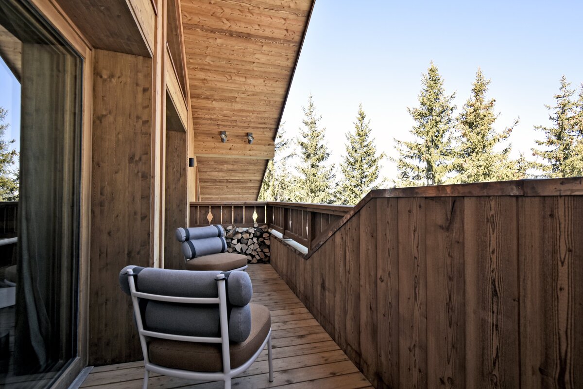 A rustic wooden chalet balcony with two modern chairs, stacked firewood, and pine trees under a clear sky.