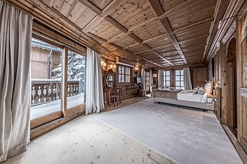 A warm, wood-paneled bedroom with a light floor and rug, featuring large windows that reveal a snowy balcony and winter scenery.
