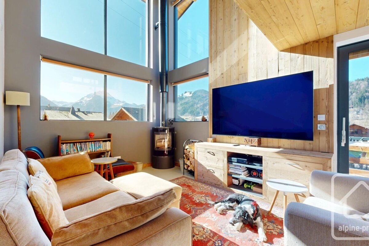 Bright modern living room with floor-to-ceiling windows showing mountain views. Features a large TV, wood stove, comfy sofa, and a dog on the rug.