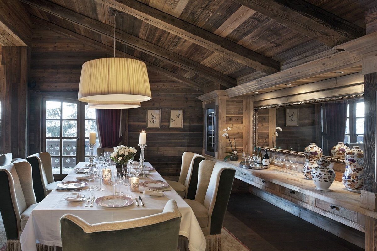 A cozy, luxurious chalet dining room with rich wooden walls/ceiling, a long table set for a meal, elegant lighting, and large windows.