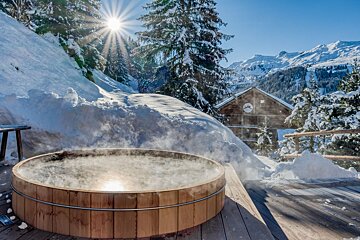 A steaming wooden hot tub on a sunny, snowy deck, with a mountain chalet, snow-covered pine trees, and distant peaks under a clear sky.