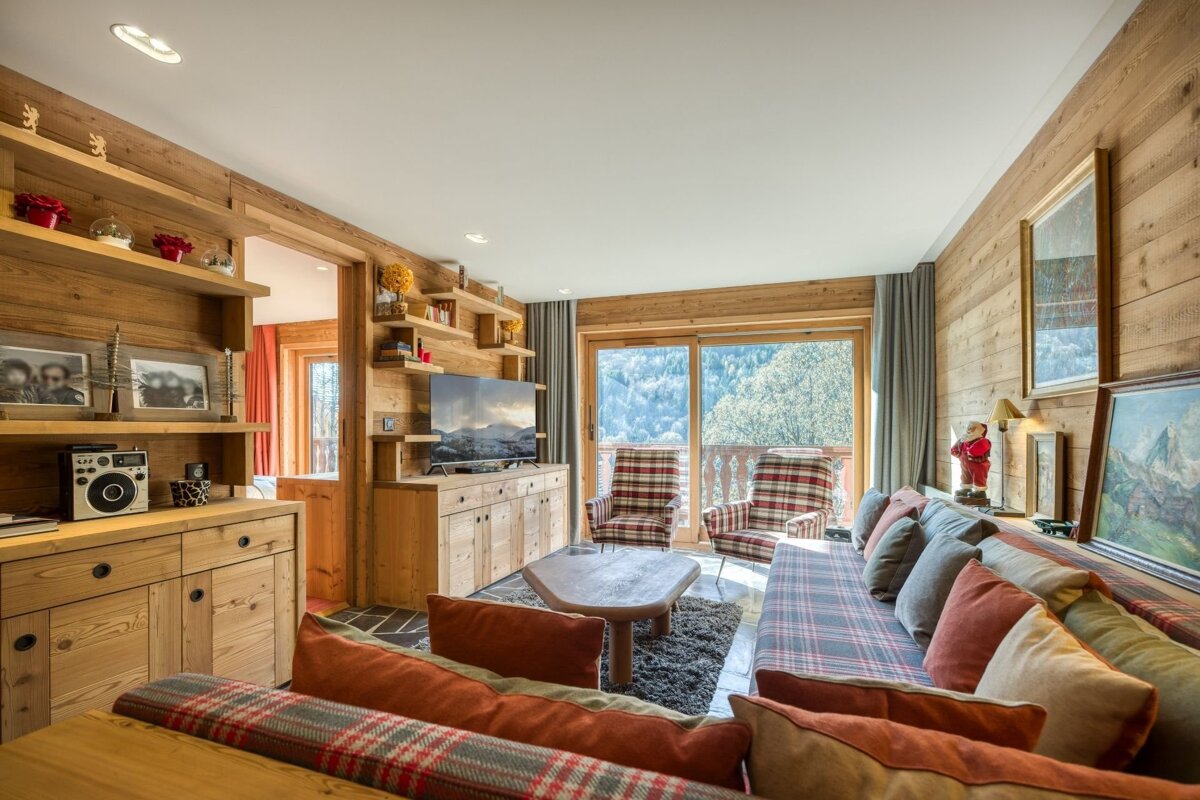 A cozy, wood-paneled chalet living room with plaid sofas and armchairs, overlooking a mountain view from a large balcony window. Warm and rustic decor fills the space.