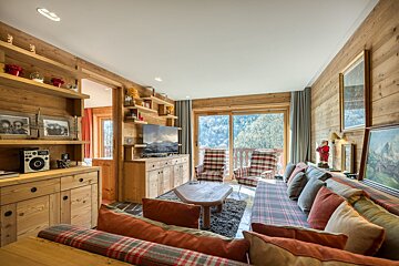 A cozy, wood-paneled chalet living room with plaid sofas and armchairs, overlooking a mountain view from a large balcony window. Warm and rustic decor fills the space.
