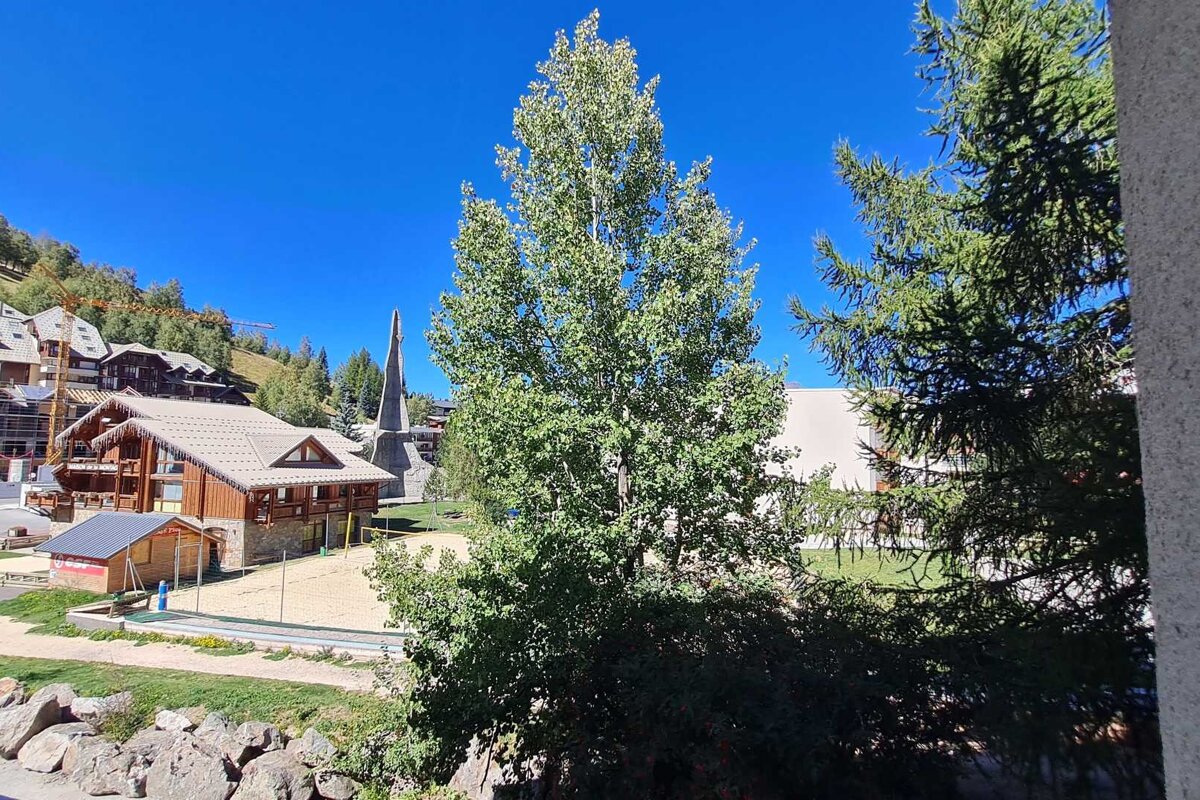 Sunny alpine village view featuring traditional chalets, a sandy court, a monument, and lush green trees against a clear blue sky.