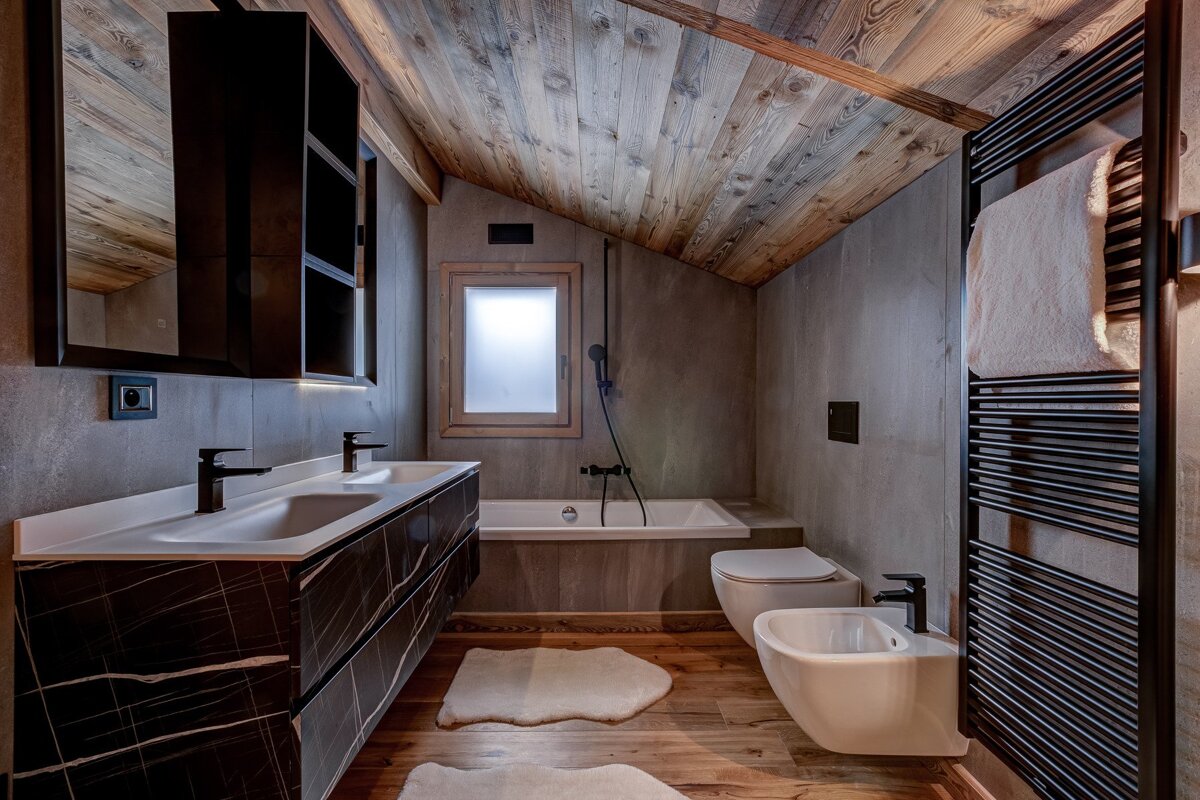 A modern, stylish bathroom features a slanted wooden ceiling, concrete walls, a dark marble vanity, black fixtures, and a freestanding tub.