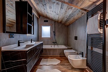 A modern, stylish bathroom features a slanted wooden ceiling, concrete walls, a dark marble vanity, black fixtures, and a freestanding tub.