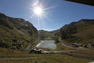 Bright sun illuminates a serene mountain valley with a sparkling lake reflecting the light, surrounded by green slopes and clear blue sky.