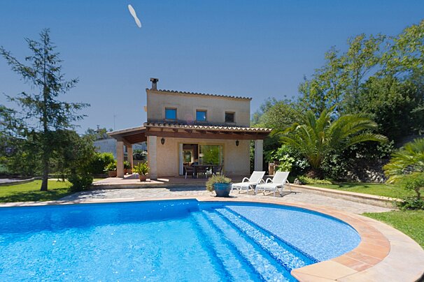 A charming villa with a sparkling blue swimming pool, surrounded by lush green foliage under a clear, sunny sky. Sun loungers await beside the pool.