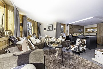 Cozy, rustic-modern chalet living room with L-shaped sofa, cowhide patterns, shaggy rug, and wood paneling, open to a dark, sleek kitchen.
