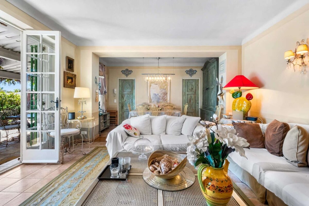 A bright, eclectic living room features white sofas, colorful decor, and a dining area. French doors open to an inviting outdoor patio.