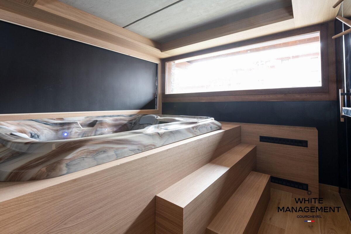 A modern indoor hot tub built into a light wood platform with steps, featuring dark walls and a bright window.