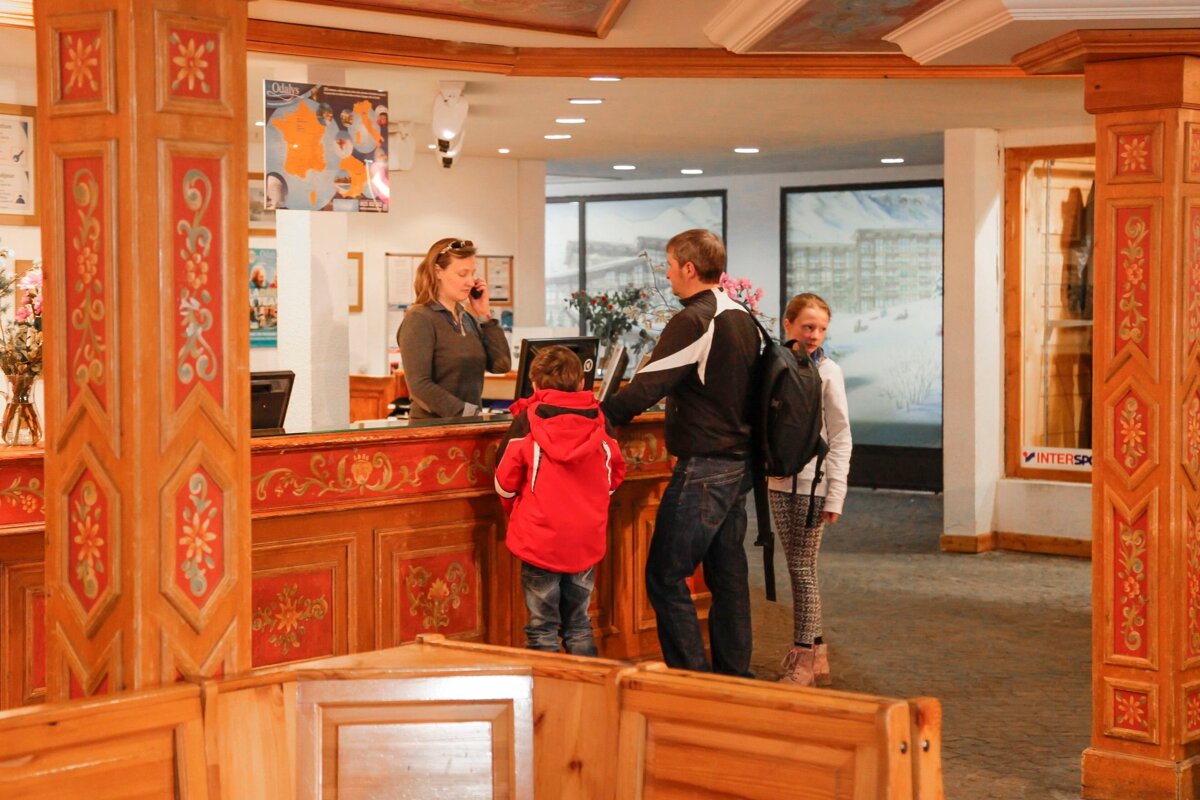 A family with two kids checks in at a rustic ski lodge reception. A female receptionist is on the phone. Ornate wooden and red decor.
