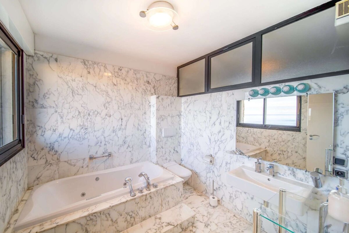 A luxurious bathroom showcasing white marble walls and floor, featuring a built-in bathtub, toilet, sink, large mirror, and windows.
