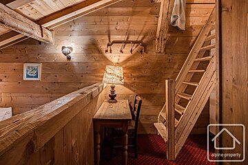 A rustic, wood-paneled room featuring a wooden desk and chair, a small table lamp, and wooden stairs leading upward.
