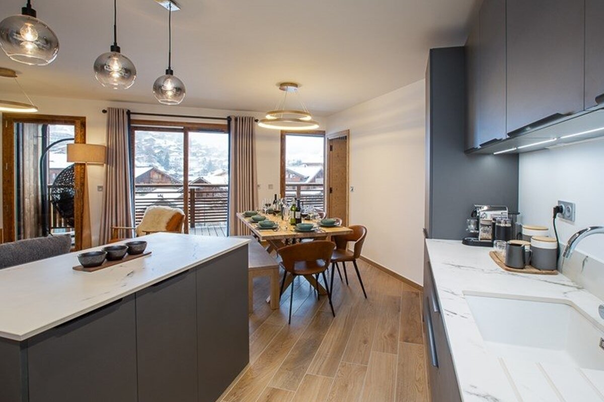 Modern kitchen and dining area in a chalet, featuring sleek grey cabinets, wooden floors, and snowy mountain views.