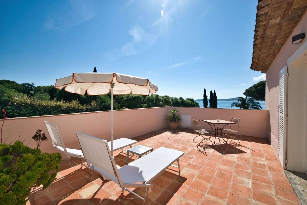 Bright, tiled terrace with white loungers, umbrella, and a small table, offering a panoramic view of the sparkling blue sea and green landscape.