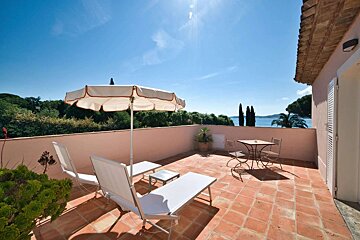 Sunny terracotta terrace with loungers, umbrella, and a dining set, overlooking a sparkling blue sea and distant green trees.