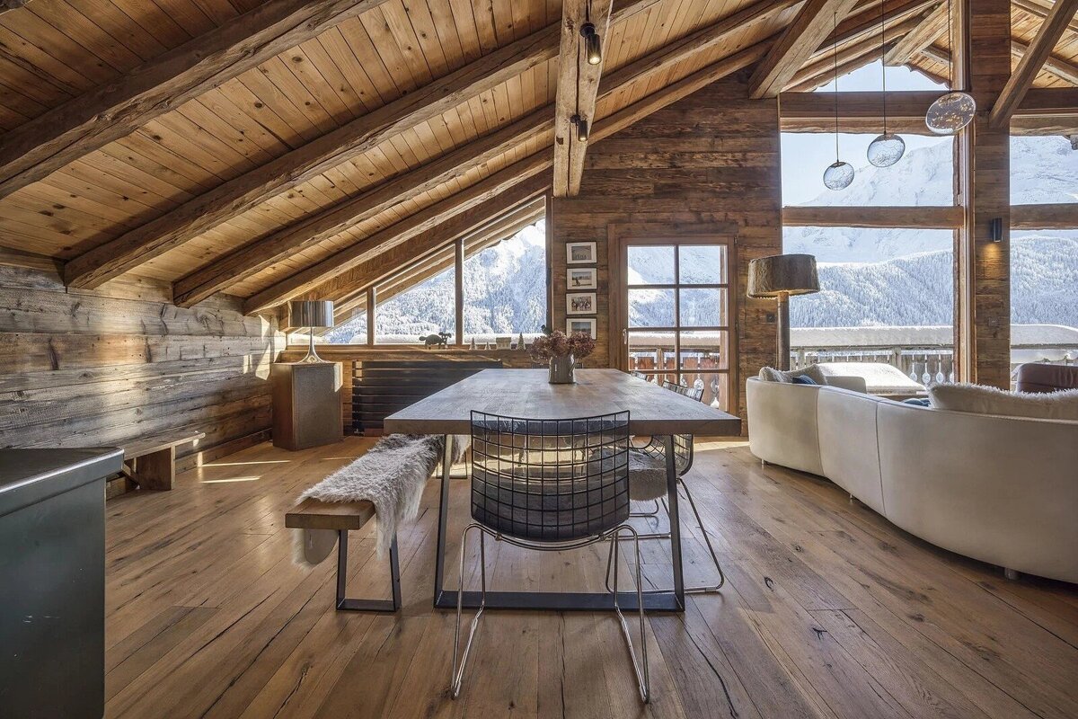 Rustic wooden chalet room with a dining table, modern furniture, and panoramic windows showing snowy mountain peaks.