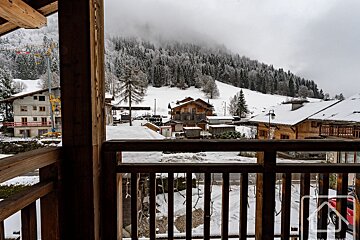 Winter view from a wooden balcony of a snowy alpine village nestled amidst snow-covered forests under an overcast sky.