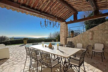 Outdoor covered dining area with a long table, chandelier, and a scenic view of a pool and distant landscape under a clear blue sky.