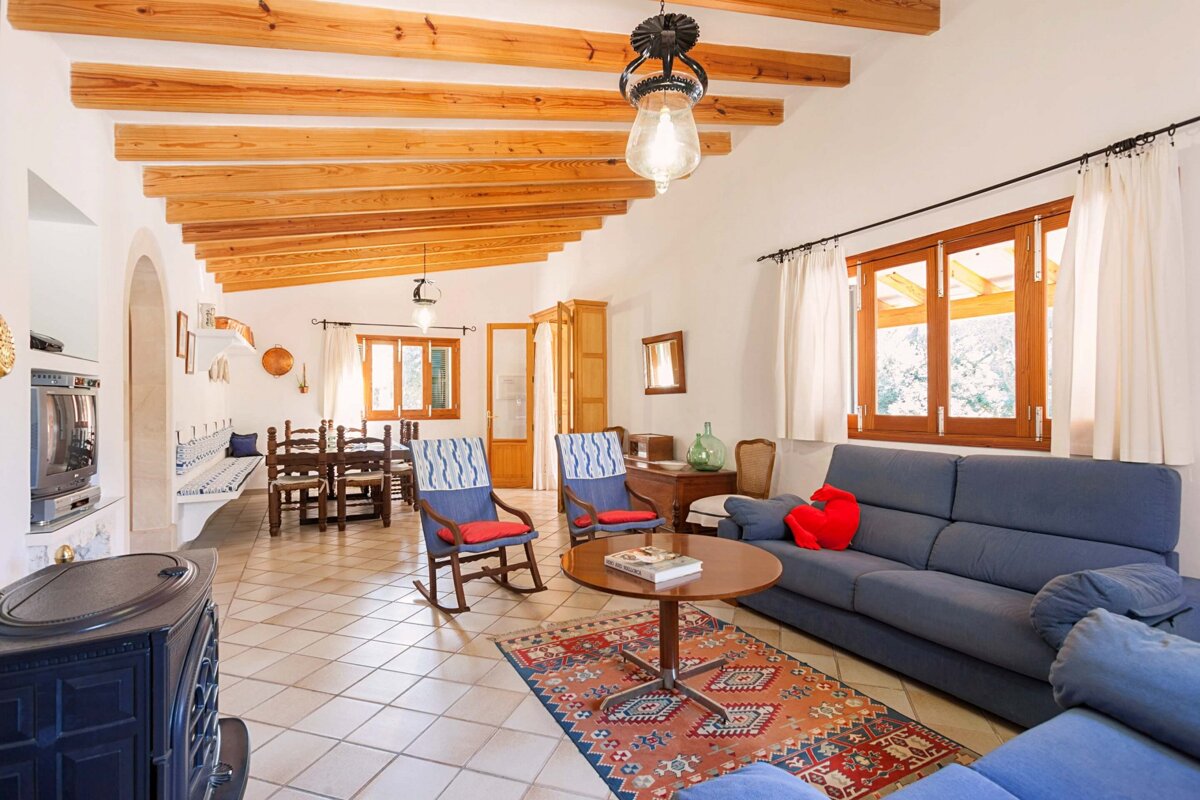 A spacious, rustic open-plan living/dining room features exposed wooden beams, tiled floors, blue sofas, a colorful rug, a wooden dining set, and a stove.