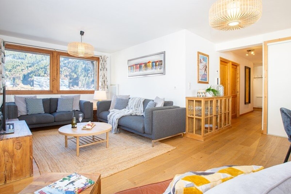 Cozy, bright living room featuring grey sofas, a round wooden coffee table, large windows with mountain views, and warm wooden flooring.