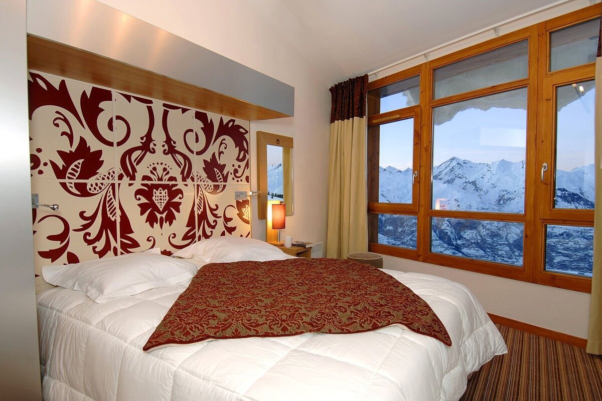 A stylish bedroom with a decorative red headboard, featuring a large window overlooking majestic snow-capped mountains.