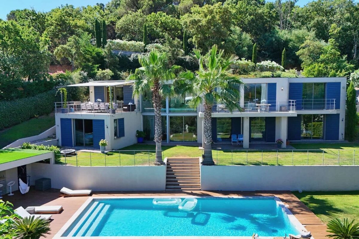 A modern multi-level house with blue shutters, balconies, and palm trees, set against a lush green hillside, with a large swimming pool and deck.