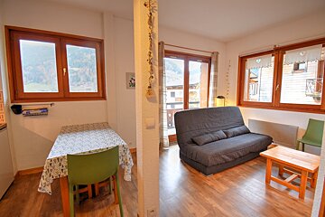 Bright, cozy room with a grey futon, dining table, and large windows revealing mountain and village views. Wooden floors.