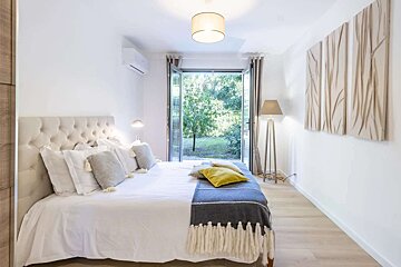 Bright, modern white bedroom with a large bed, tufted headboard, and open glass doors revealing a lush green garden. Clean and inviting.