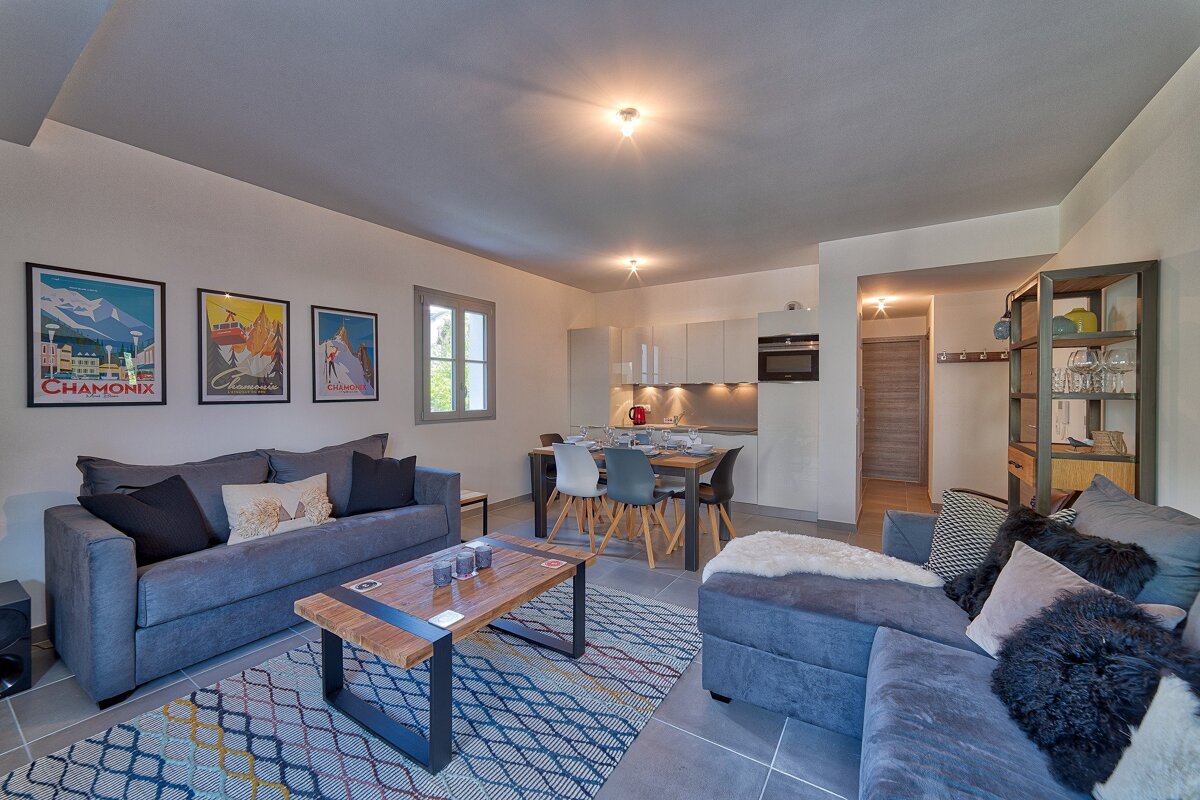 Stylish open-plan apartment interior with gray sofas, patterned rug, wooden coffee table, Chamonix art, and a connected dining area and modern kitchen.