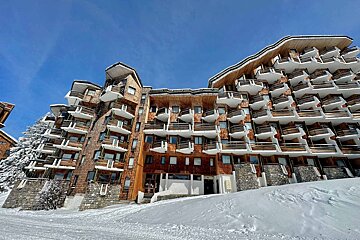 A grand wooden chalet-style building with numerous balconies stands in a snowy mountain landscape under a clear blue sky, evoking a winter resort scene.