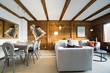 Cozy cabin interior featuring wood-paneled walls, exposed ceiling beams, a large grey sectional sofa, and a dining set.