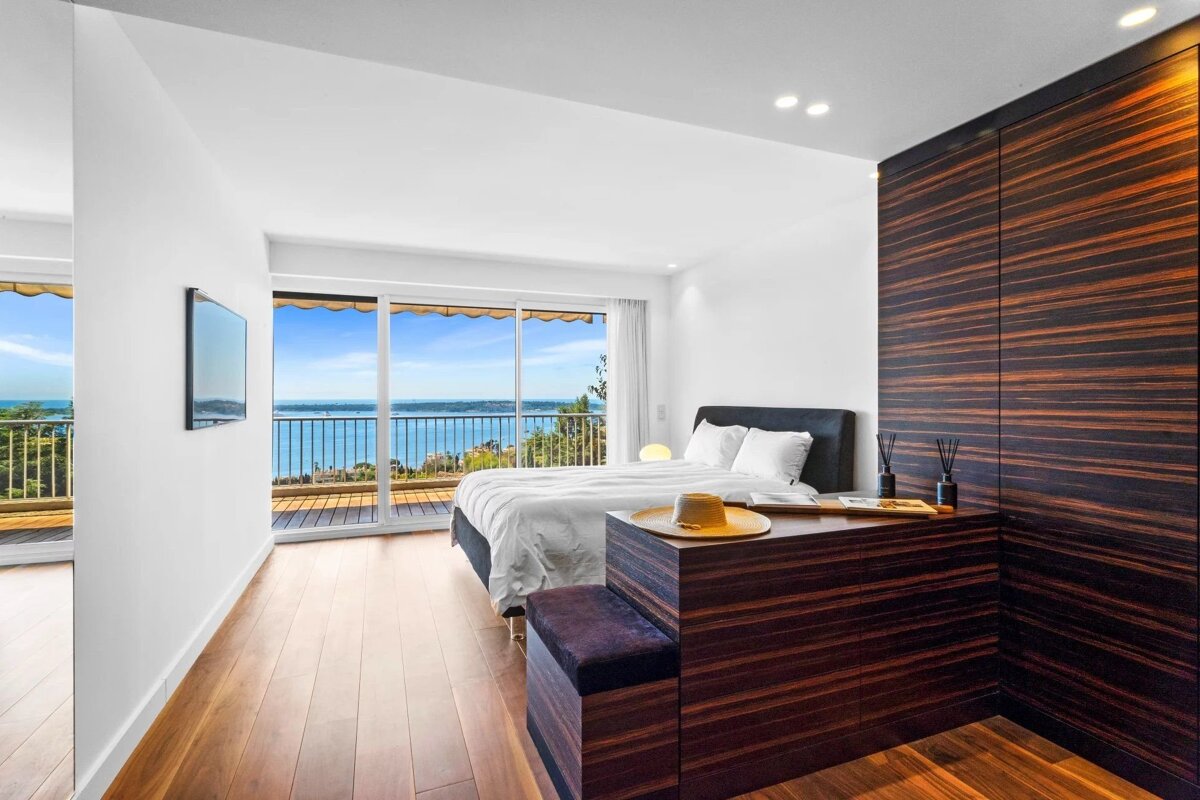 A modern bedroom with white walls, wooden details, and large sliding doors opening to a balcony with sea views.