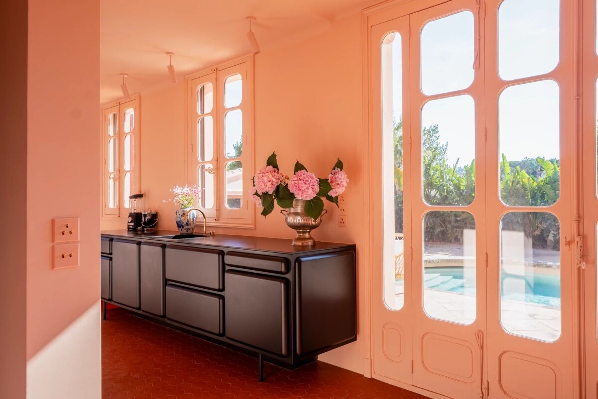 A peach-colored kitchen with dark cabinets, pink hydrangeas, and tall glass doors opening onto a sunny backyard pool.