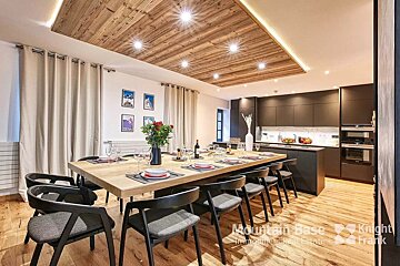 A stylish open-plan dining room and modern kitchen. Features a long wooden table, dark cabinets, hardwood floors, and a warm wooden ceiling with lights.