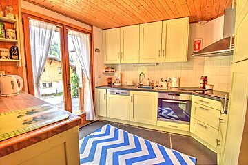 A cozy kitchen with light yellow cabinets, wooden ceiling, and a blue/white chevron rug. French doors open to an outdoor view with a kettle on the counter.