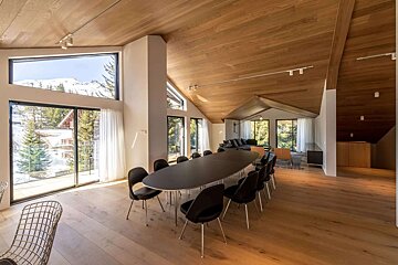 Spacious, modern dining room with wood ceilings, a long table, and large windows revealing a stunning snowy mountain landscape.