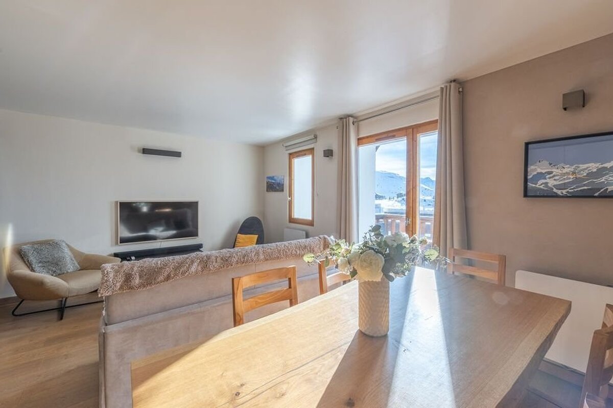 A bright, cozy living and dining space with wooden furniture and glass balcony doors opening to a scenic, snowy mountain view.
