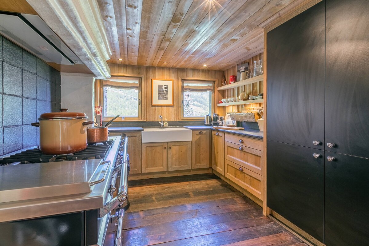 Cozy chalet kitchen with wood-paneled walls and ceiling, featuring a professional stove, wooden cabinets, and large black storage doors.