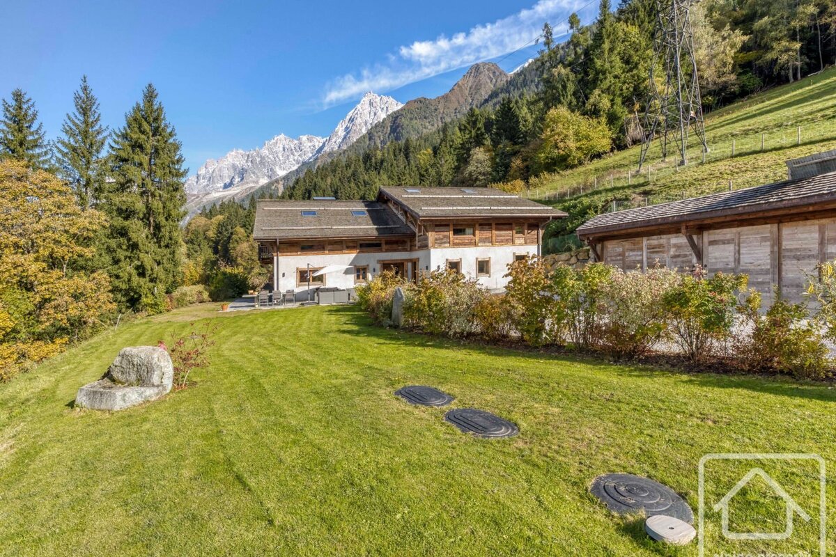 A rustic chalet with a green lawn sits before majestic snow-capped mountains and evergreen forests under a clear blue sky.